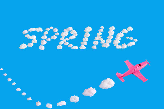 Pink airplane goes into turn with clouds trail on blue background and word spring of abstract shapes of cloud. Minimal composition. 