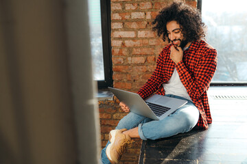  A curly-haired student with an unusual haircut sits at the university with a laptop. african in red shirt and jeans