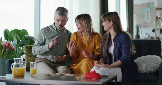 Happy candid family trio of mother, father and daughter playing domino table game enjoying relaxing leisure activity at home apartment.