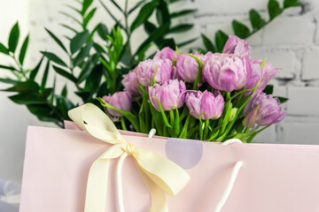 Close-up detail of fresh beautiful present of tender violet peony tulips bouquet and paper gift bag with romantic bow. Wedding or anniversary celebration surprise background. Pastel floral decoration