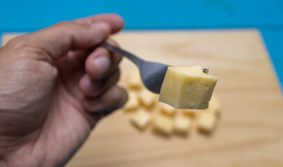 Yellow cheese cut into cubes on a cutting board on a blue table and a metal fork. Snack used to celebrate at home with family and friends.