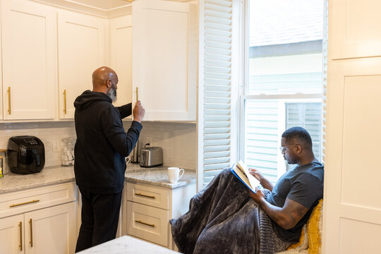 Man reading in nook at home while partner fixes coffee drink, gay couple