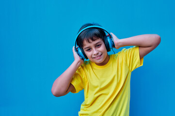 Portrait of smiling boy in yellow t-shirt and blue headphones listening to music, on blue background. Copy space