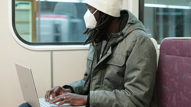 Afro man in mask uses laptop in the passenger seat on the train. Hands pushing buttons, doing urgent work. Waiting for the train to depart.