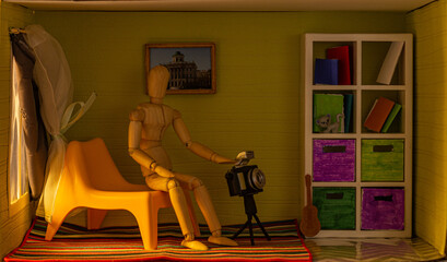 Fototapeta na wymiar Figurine of a puppet man sitting in a chair by the window in a toy room with a camera