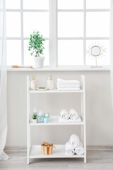 Fototapeta na wymiar Shelf unit with cosmetics and towels near window in bathroom