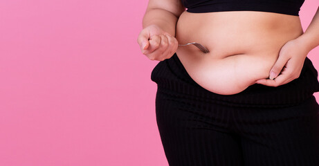 Obesity unhealthy weight loss concept. Hand of fat woman holding fork on her belly while standing over isolated pink background. Chubby overweight hold stomach waistline want start dieting.