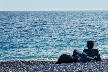 Fototapeta premium A man lies in the evening on a pebble beach in the city of Nice in France and looks at the water of the Mediterranean Sea.