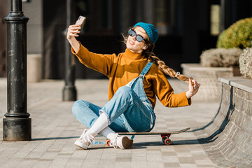 Young stylish caucasian woman skateboarder in a hat, sunglasses and jeans is using a phone next to a skate board in the city. Theme of active youth recreation in an urban environment