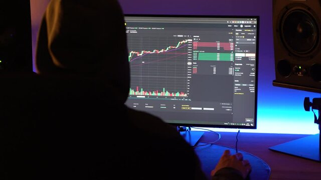 Backview of a trader with hoodie looking at financial chart trading bitcoin and cryptocurrency in the dark