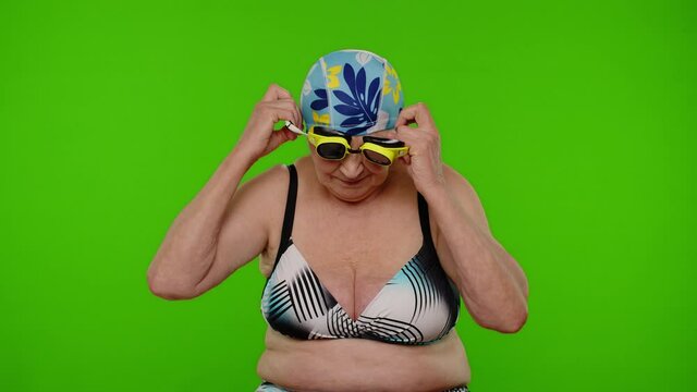 Senior woman tourist wearing swimming glasses, looking at camera, dancing on chroma key background. Travel, summer holiday vacation, beach party. Elderly grandmother in swimsuit, indoor studio shoot