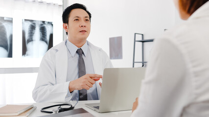 Fototapeta premium Serious Asia male doctor in white medical uniform using computer laptop is delivering great news talk discuss results or symptoms with female patient sit at desk in health clinic or hospital office.