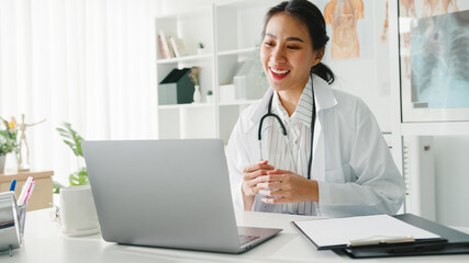 Young Asia lady doctor in white medical uniform with stethoscope using computer laptop talking video conference call with patient at desk in health clinic or hospital. Consulting and therapy concept.