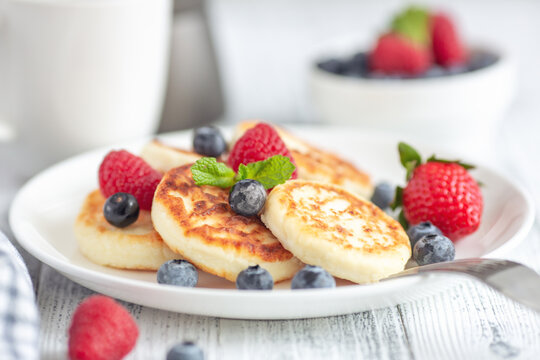 Close up of cottage cheese pancakes with fresh berries on white wooden table. Curd fritters with berries, syrniki. Soft focus - Image