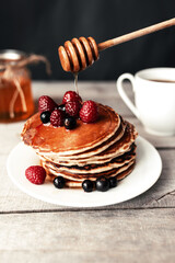 Pancakes with berries and honey on a white plate, wooden spoon, jar, coffee cup