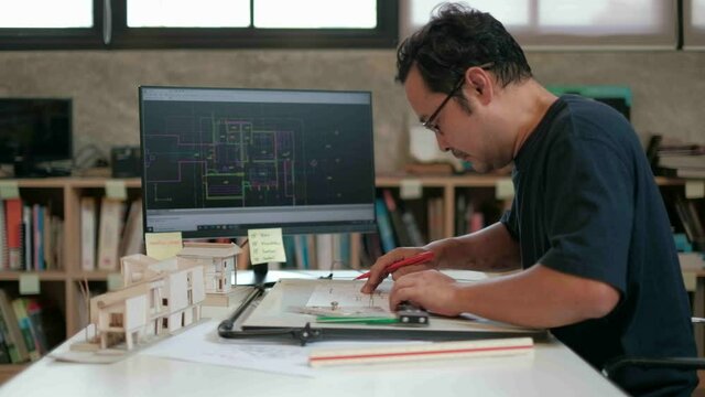 An Asian architect in a dark shirt is working on a design in his studio using a computer-aided design program and drafting board to drafting the construction plan.
