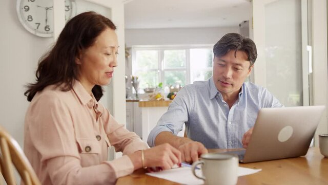 Mature Asian couple sitting at table at home using laptop to organise household bills and finances - shot in slow motion