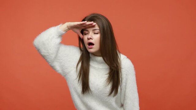 Focused attentive woman with brown hair in fluffy white sweater looking far away with hand over head, searching, expecting to see something at horizon. Indoor studio shot isolated on orange background