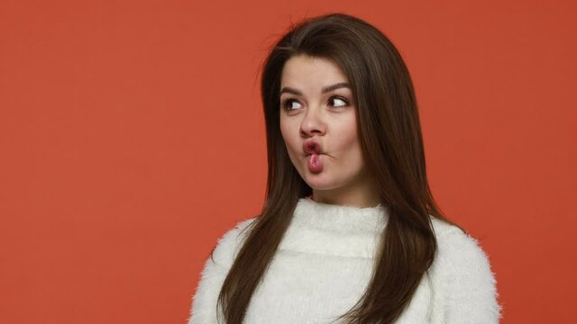 Funny ridiculous brunette woman in fluffy white sweater putting lips like fish and trying to talk, having fun, fooling around making silly comic face. Indoor studio shot isolated on orange background