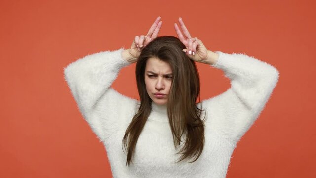 Irritated woman in white sweater showing bull horns holding fingers on head making threatening grimace to camera expressing anger and danger. Indoor studio shot isolated on orange background
