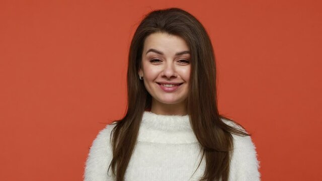 Funny brunette woman in white sweater crossing eyes making silly dumb face, having fun grimacing demonstrating dumb ridiculous face expressions. Indoor studio shot isolated on orange background