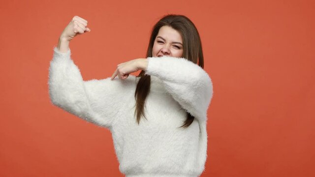 I am strong and independent. Happy positive and successful woman in fluffy white sweater showing her arm muscles raising hands up, bragging with power. Indoor studio shot isolated on orange background