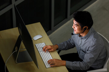 Top view portrait focus to asian man call center wearing headphone sitting and taking with customer feel happy on smiling and typing on keyboard computer monitor