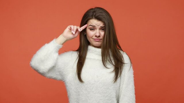 Upset frustrated brunette woman in white fluffy sweater twirling finger to temple showing cuckoo stupid gesture, looking at camera with sad expression. Indoor studio shot isolated on orange background