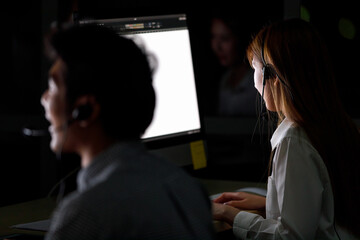 Shooting photo from back focus to asian woman call center as sitting near man staff. Woman helpdesk doing look at computer monitor and typing keyboard in office at night