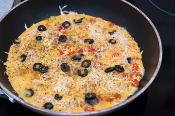 Raw omelet in a round frying pan, top view. Eggs, milk, cheese, olives, tomatoes, spices. Ready for further cooking. 
