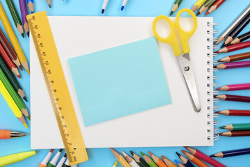 On a blue background among colored pencils there is a notebook on top of which is a clean blue sheet and yellow scissors with a ruler. The concept of training. Template. Space for text