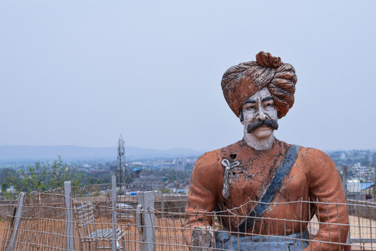 Front View Of 30-40 Years Old Indian Warrior Statue, Wearing Orange Color Turban And Indian Traditional Cloths Standing Still In A Sunny Day At Kolhapur City Maharashtra India On Blur Background 