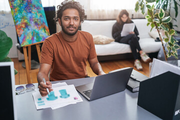 Portrait of a male indian designer and stylist he does his work sitting at table in background of his colleague she sits on sofa.