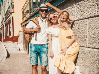 Group of young three stylish friends posing in the street. Fashion man and two cute female dressed in casual summer clothes. Smiling models having fun in sunglasses.Cheerful women and guy outdoors