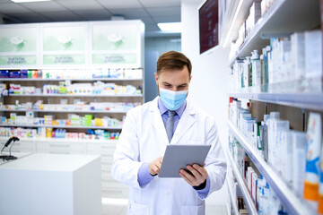 Pharmacist wearing face mask and white coat working in pharmacy store on tablet computer during...