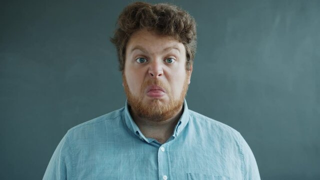 Portrait of guy showing tongue with hostile face then putting on glasses and looking at camera standing on gray background. Expressions and grimaces concept.