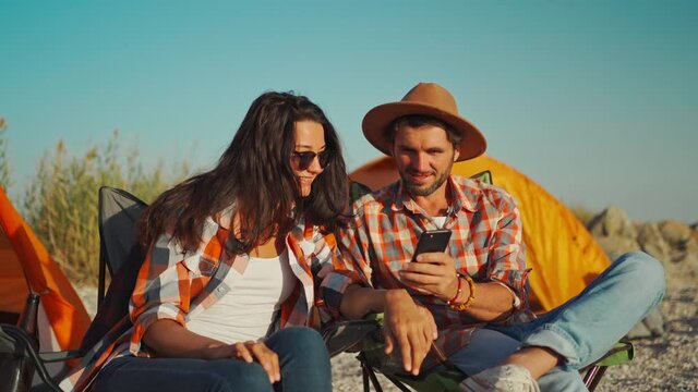 beautyful romantic couple in checkered shirts sitting at seaside in camp chairs and looking photos and videos on smartphone. summer vacation and travel to sea with tents, looking memories on device