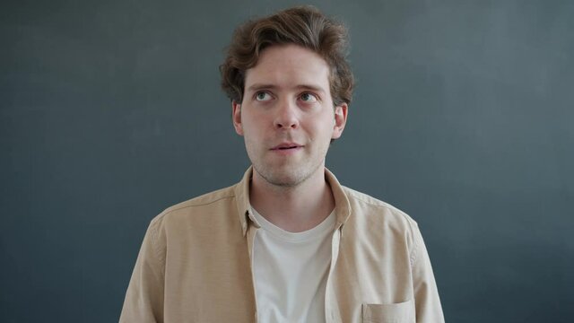 Frustrated guy is rotating eyes looking around then staring at camera with confused face on gray background. Emotions and body language concept.
