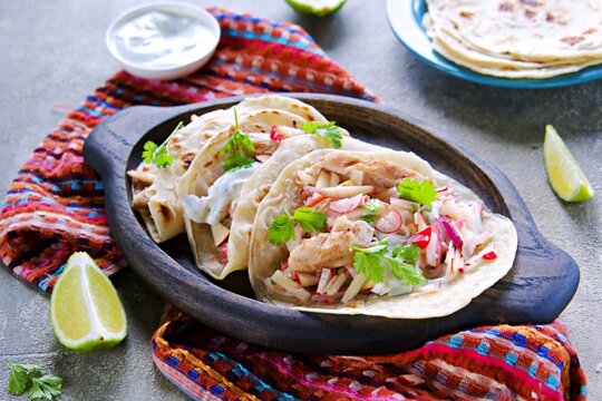 Tacos with chicken, radish and apple salad and sour cream sauce in a round wheat tortilla on an oval wooden plate on a gray concrete background.