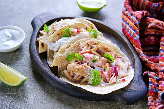 Tacos with chicken, radish and apple salad and sour cream sauce in a round wheat tortilla on an oval wooden plate on a gray concrete background.