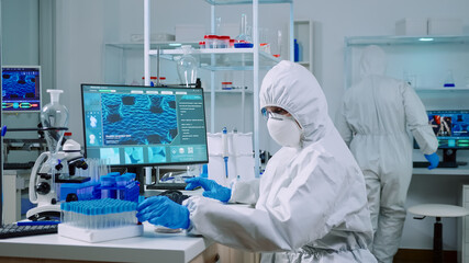 Doctor with protection suit working with blood samples in laboratory typing on computer. Chemist examining vaccine evolution using high tech for research in treatment development against covid19 virus