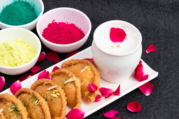 Holi Celebration Theme - Gujia Or Gujiya Mithai Made In Desi Ghee Pista Badam Served With Cold Thandai Bhang. Red Green Yellow Colorful Gulal And Rose Flower Petal Decoration On Black Background