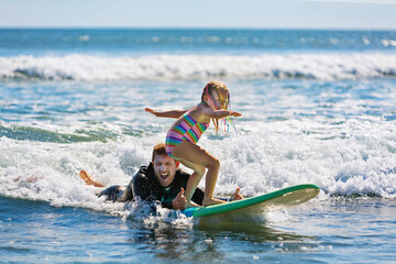 Little surf girl - young surfer learn to ride on surfboard with instructor at surfing school. Active family lifestyle, kids water sport lessons, swimming activity in summer camp. Vacation with child.