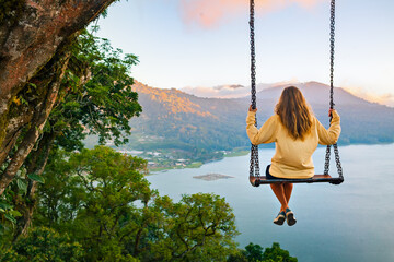Summer vacation. Young woman sit on tree rope swing on high cliff above tropical lake. Happy girl looking at amazing jungle view. Buyan lake is popular travel destinations in Bali island, Indonesia - obrazy, fototapety, plakaty