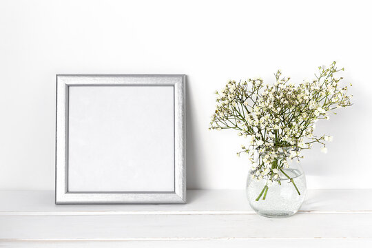 White spring flowers in a glass vase with a silver photo frame on a white table. Cozy white interior