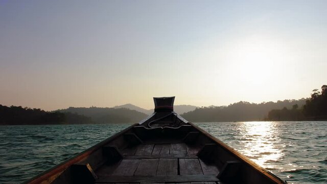 Taking a Wooden traditional thai longtail boat ride at a tropical Cheow Lan lake at sunset in Khao Sok National Park, Thailand in one of the famous iconic Thai longtail boats in slow motion