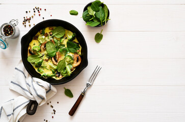 Omelet with spinach and mushrooms in a cast-iron pan on a light wood background. Fritata with...