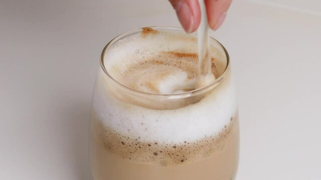 Person hand stirring latte coffee with glass spoon. Making morning coffee with milk, stirs sugar in a cup.