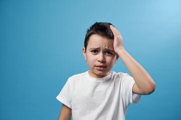 Boy holding his temple. Facial emotion on blue background with copy space