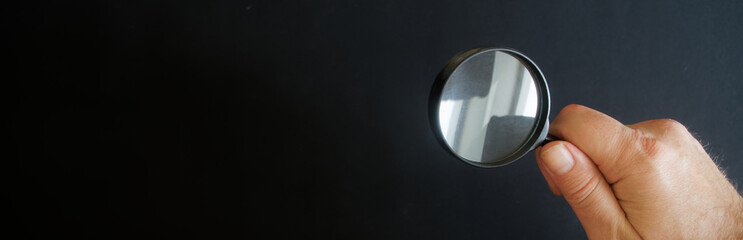 A hairy hand of an adult man holds a glass magnifier on a black background. Search, investigation...